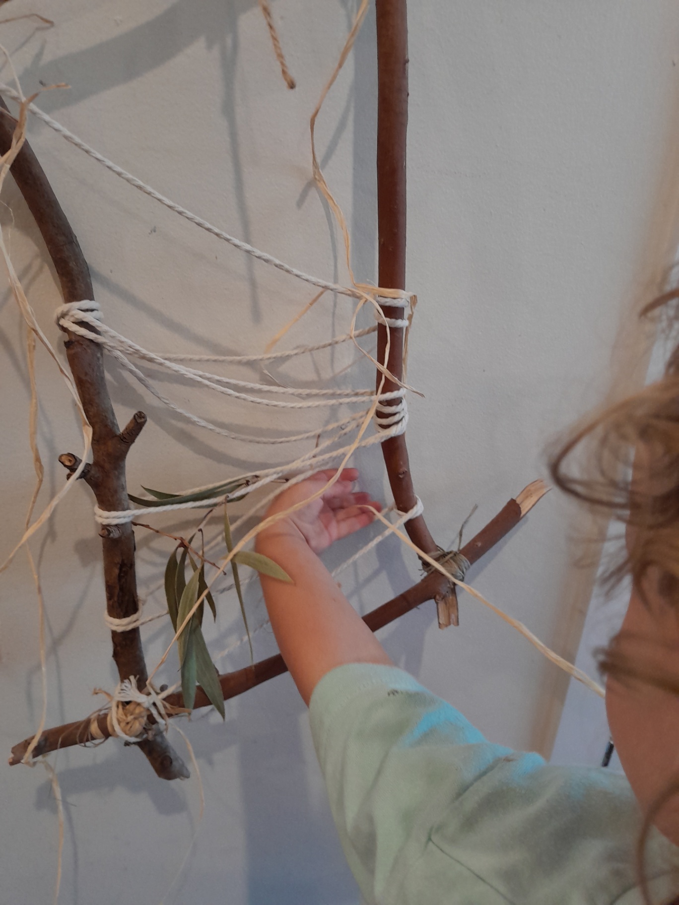 Tiny hands weaving raffia and eucalyptus through a stick loom